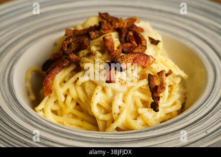 Eine wunderschön überzogene Portion Spaghetti Carbonara mit knusprigem Pancetta auf einer gestreiften Keramikplatte. Die cremige Soße und die goldenen Töne erzeugen einen Stockfoto