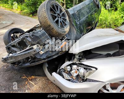 Silberne Limousine, die über schwarze Fahrzeuge gekippt wurde, verheddern sich nach einem Zusammenstoß auf ruhiger, bewaldeter Straße. Stockfoto