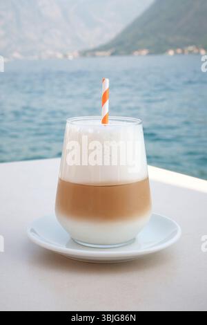 Glas Eislatte mit Orangenstroh und cremigem Milchschaum auf einem weißen Tisch am Meer. Hintergrund der Küstenberge Stockfoto