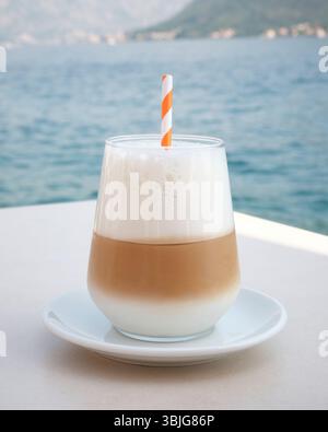 Eislatte mit dickem Schaum und gestreiftem Stroh auf einer weißen Untertasse, serviert auf einem Tisch am Meer mit bergiger Küste Stockfoto