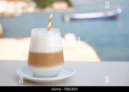 Glas Eislatte mit Milchschaum und Stroh auf einem Tisch am Meer. Verschwommenes Boot und Küstengebäude im Hintergrund Stockfoto