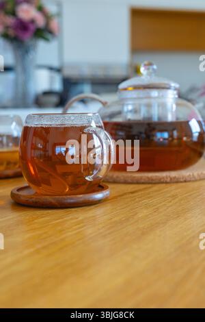 Teekanne aus Glas und Tassen mit heißem Tee und Zitrone auf Holztisch im hellen Wohnzimmer. Warmes Licht und gemütliches Interieur. Konzept von Wellness und Entspannung Stockfoto