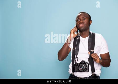 Junger schwarzer Mann mit einer DSLR-Kamera um den Hals, der auf seinem Mobilgerät spricht. Ein Fotograf trägt Hosenträger und steht selbstbewusst im Studio, während er mit einem Kunden auf seinem Handy spricht. Stockfoto