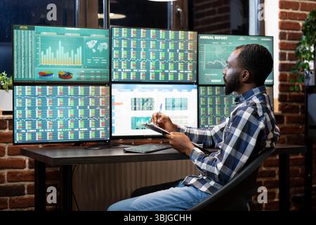 Der afroamerikanische Forex-Trader untersucht Live-Marktdaten auf mehreren Monitoren und macht sich Notizen in seinem Clipboard in einem modernen Arbeitsbereich. Er konzentriert sich während einer Handelssitzung auf einer technisch versierten Workstation. Stockfoto