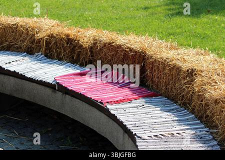 Eine lange, geschwungene Parkbank ist mit gestreiften Teppichen bedeckt und von einer Reihe Heuballen auf Kopfsteinpflasterpflaster mit grünem Gras überzogen Stockfoto