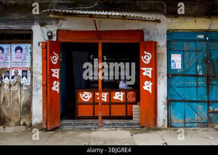 Ein kleines Medizingeschäft in Jessore, Bangladesch, mit bengalischen Schildern, orangefarbenen Fensterläden und einem Mann, der hinter einer Apotheke sitzt. Stockfoto