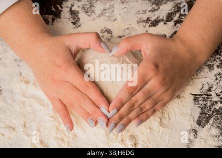 Kochen mit Liebe. Weibliche Hände, die den Teig in Herzform halten. Berühren Sie den rohen Teig sanft mit den Händen in Form eines Herzens. Weibliche Hände, die Teig halten Stockfoto