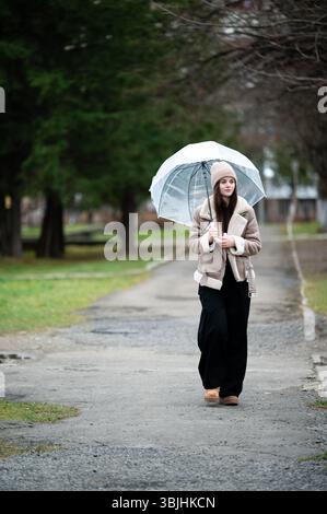 Eine modische junge Frau schlendert einen üppigen Parkweg hinunter und hält einen transparenten Regenschirm, warm gekleidet für einen kühlen und luftigen Nachmittag in der Natur. Stockfoto