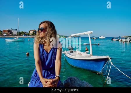 Frau in blauem Kleid sitzt auf Felsen in der Nähe des Meeres mit Booten auf türkisfarbenem Wasser. Sommerurlaub und Küstenleben Konzept Stockfoto