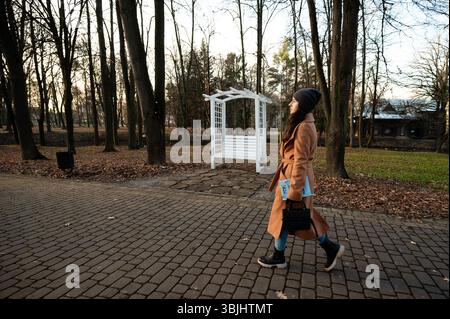 An einem frischen Nachmittag spaziert eine junge Frau in einem warmen Mantel und einem stilvollen Hut durch einen ruhigen Park, umgeben von hohen Bäumen und einem malerischen Bogengang Stockfoto