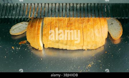 Frisch Gebackener Brotbrot Wird In Scheiben Geschnitten. Eine Nahaufnahme eines frisch gebackenen Brotlaibs, der von einer Brotschneidemaschine geschnitten wird, mit verstreuten Krümeln. Stockfoto