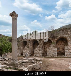 Eine alte Steinsäule steht in der Nähe eines Gebäudes mit Bögen in Ephesus, Türkei. Felsen umgeben die Ruinen. Der Himmel darüber ist blau mit flauschigen weißen Wolken Stockfoto