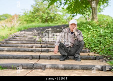 In einem üppig grünen Park an einem sonnigen Maitag sitzt ein Japaner Ende 70 mit kariertem Hemd und weißem Hut auf der Treppe. Umgeben von spr Stockfoto