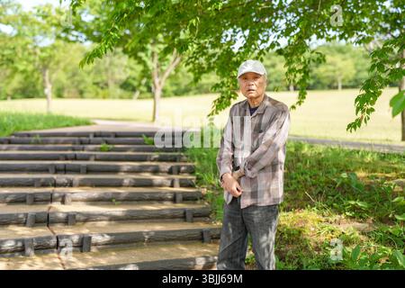 In einem üppig grünen Park an einem sonnigen Maitag steht ein Japaner Ende 70 mit kariertem Hemd und weißem Hut still auf Stufen. Umgeben Stockfoto
