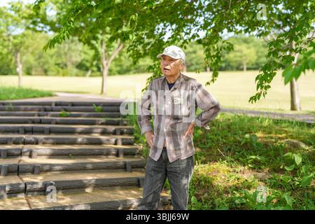 In einem üppig grünen Park an einem sonnigen Maitag steht ein Japaner Ende 70 mit kariertem Hemd und weißem Hut still auf Stufen. Umgeben Stockfoto