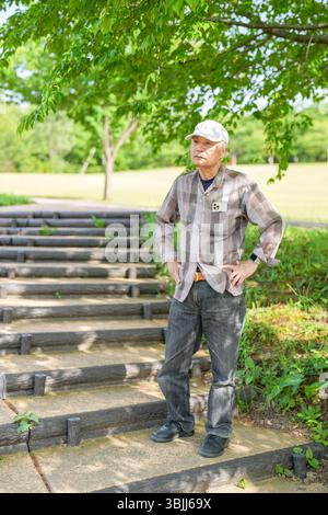 In einem üppig grünen Park an einem sonnigen Maitag steht ein Japaner Ende 70 mit kariertem Hemd und weißem Hut still auf Stufen. Umgeben Stockfoto