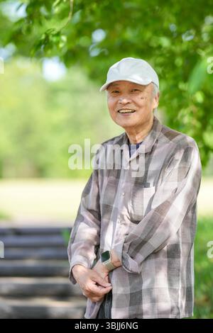 In einem üppig grünen Park an einem sonnigen Maitag steht ein Japaner Ende 70 mit kariertem Hemd und weißem Hut still auf Stufen. Umgeben Stockfoto