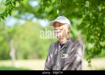 In einem üppig grünen Park an einem sonnigen Maitag steht ein Japaner Ende 70 mit kariertem Hemd und weißem Hut still auf Stufen. Umgeben Stockfoto