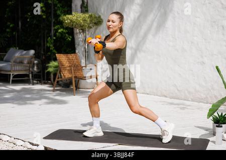 Fit Frau in Aktivkleidung, die mit orangen Kurzhanteln und Handgelenkgewichten im Freien trainieren, Stärke, Fokus und gesunde Lebensweise demonstrieren. Stockfoto