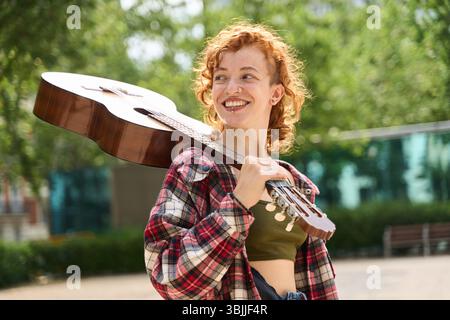 Porträt einer jungen rothaarigen Straßenkünstlerin, die eine Gitarre auf der Schulter trägt, wegblickt und lächelt, in einem Park Stockfoto