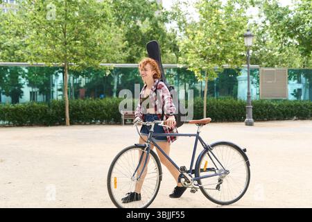 Junge rothaarige Musikerin, die mit Fahrrad und Gitarre in einem Stadtpark spaziert und einen sonnigen Sommertag genießt Stockfoto