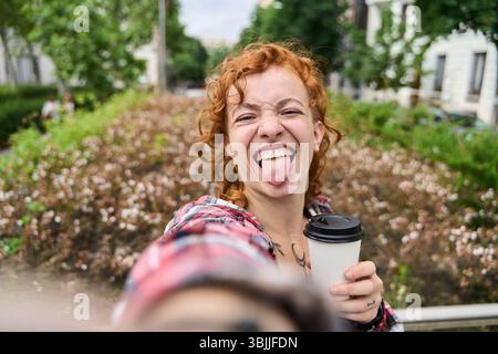 Junge rothaarige Frau mit lockigen Haaren, Nasenpiercing und Tätowierungen, trägt ein kariertes Hemd, macht ein Selfie, indem sie ihre Zunge herauszieht, während sie einen Tak hält Stockfoto