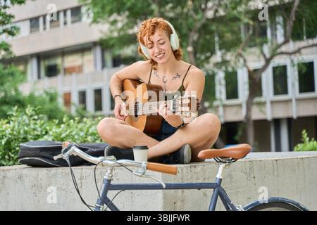 Rothaarige Frau mit Tattoos und Kopfhörern, die gerne Akustikgitarre im Freien spielt, neben ihrem Fahrrad mit Gitarrenkoffer und Kaffee zum Mitnehmen sitzt Stockfoto