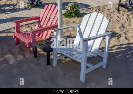 Zwei bunte adirondack-Stühle und ein kleiner Beistelltisch sitzen am Sandstrand und sorgen für eine entspannende Szene Stockfoto