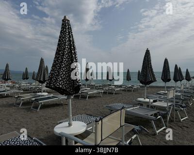 Geschlossene Sonnenschirme und Liegestühle am Morgenstrand vor dem Hintergrund der Küste Stockfoto
