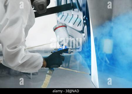 Professioneller männlicher Autolackierer sprüht Autoteile in der Garage mit dem Airbrush auf blaue Farbe. Der Mann arbeitet mit der Spritzpistole. Stockfoto