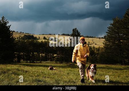 Frau in einer gelben Jacke und ihr australischer Schäferhund, der über eine Wiese mit Wald und Hügeln unter einem stimmungsvollen Himmel in Zlatibor, Serbien, spaziert. Weiblich Stockfoto