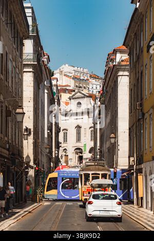 Klassische gelbe Straßenbahn und moderne Straßenbahn fahren durch eine enge, sonnendurchflutete Straße mit alten Gebäuden in Lissabon, Portugal - 11. Juni 2025 Stockfoto