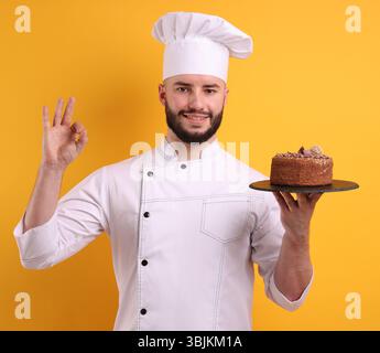 Glücklicher Konditor in Uniform, der Schokoladenkuchen hält und eine gute Geste auf orangem Hintergrund zeigt Stockfoto