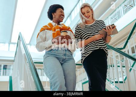 Zwei weibliche Kollegen, die in einer Büroumgebung Treppen hinuntergehen und sich leicht und freundlich unterhalten. Einer hält eine Kaffeetasse und der OT Stockfoto