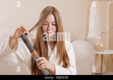 Die junge Frau stylt ihr langes, gesundes Haar zu Hause mit einem modernen Lockenstab und präsentiert eine Schönheits- und Haarpflegeroutine mit fortschrittlicher Technologie für eine Stockfoto