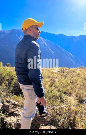 Ein Mann mit gelbem Hut und blauer Jacke steht auf einem Hügel und hält eine Kamera. Der Himmel ist klar und die Berge im Hintergrund sind majestätisch Stockfoto