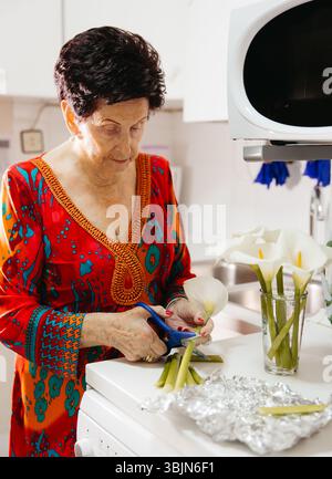 Ältere Frau, die Blumen in der Küche anordnet und Schere und Glasvase hält Stockfoto