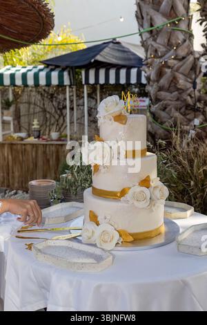 Eine atemberaubende dreistöckige weiße Hochzeitstorte mit goldenen Besätzen und weißen Rosen, bereit für die Zeremonie des Kuchens. Stockfoto
