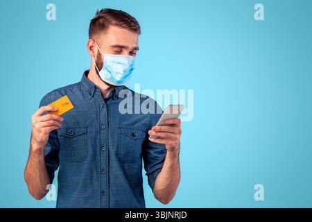 Kontaktloses Einkaufen während der Covid-Isolation. Porträt von Millennial Kerl in Gesichtsmaske hält Smartphone und Kreditkarte Stockfoto