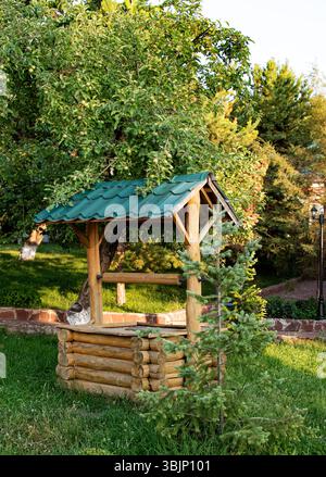 Das Foto zeigt einen dekorativen Holzbrunnen mit grünem Dach, der sich in einem Garten befindet. Umgeben von Bäumen, Gras und einem jungen Fichtenbesatz, alle eingetaucht Stockfoto