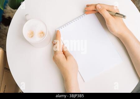 Flache Ladefläche mit Kaffee, Notizbuch und Frauenhänden, die einen Stift halten, von oben gesehen. Stockfoto