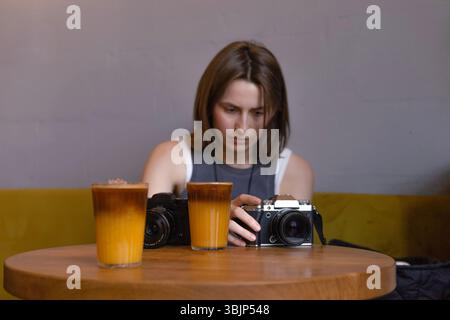 Die junge Fotografin überprüft die Kameras, während sie in einem Café mit einem Glas Kaffee und Orangensaft sitzt Stockfoto