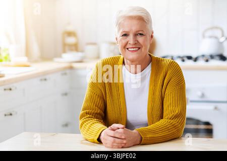 Lächelnde ältere Dame, die zu Hause am Tisch in der Küche sitzt Stockfoto