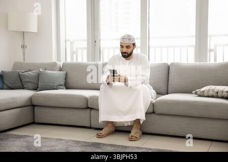 Junger Araber, der das Telefon auf der Couch im Wohnzimmer benutzt Stockfoto