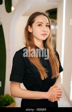 Selbstbewusste junge Krankenschwester in schwarzer Uniform stehend mit Blick auf die Kamera im modernen Klinikinnenraum, strahlt Professionalität aus Stockfoto