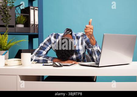 Junger schwarzer Geschäftsmann, der den Kopf auf dem Schreibtisch auflegt und die Daumen hochgibt, erschöpft, aber den Erfolg feiert. Verschlafener Büroangestellter mit Genehmigungszeichen und Nickerchen auf dem Tisch nach einem anstrengenden Arbeitstag. Stockfoto