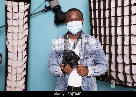 afroamerikaner hält seine DSLR-Kamera und trägt eine Gesichtsmaske, während er sich auf ein kreatives Projekt vorbereitet. Studio-Porträt des schwarzen Fotografen, der Vorsichtsmaßnahmen trifft, um sich vor Krankheiten in der Luft zu schützen. Stockfoto