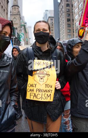Eine Person bei einem Protest hält ein gelbes Schild mit der Aufschrift „nur du kannst Faschismus verhindern“ und zeigt eine Illustration des Smokey Bear. Sie tragen einen schwarzen Regen Stockfoto