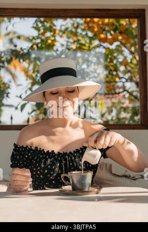 Frau mit Hut, die Milch in Kaffee am sonnigen Frühstückstisch gießt, friedlichen Moment genießt Stockfoto