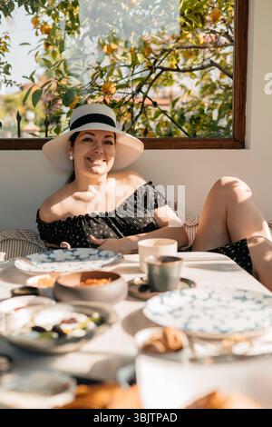 Lächelnde Frau mit Sonnenhut, die sich am türkischen Frühstückstisch auf der Terrasse mit Zitronenbaum im Hintergrund entspannt Stockfoto